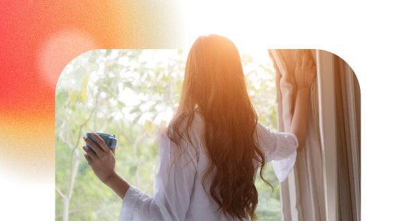 A woman with long brown hair stares out a window. She is holding a cup of coffee and resting the other hand on the windowsill.