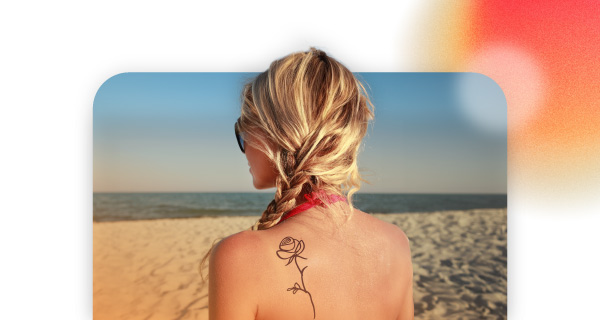 A woman with blonde hair on the beach. She has a rose tattoo on her left shoulder and is staring out at the waves.