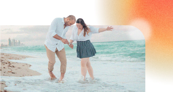 Two people, a young man and a young woman, walking through the shallow waters of the ocean. They are holding hands and chatting, looking utterly focused on each other.