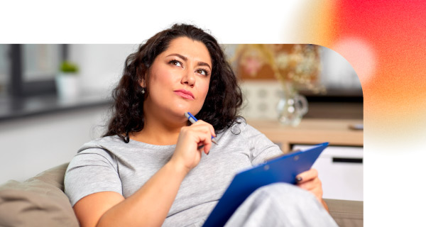 A woman sits on the couch, with a clipboard on her lap. She holds a pen to her chin thoughtfully.